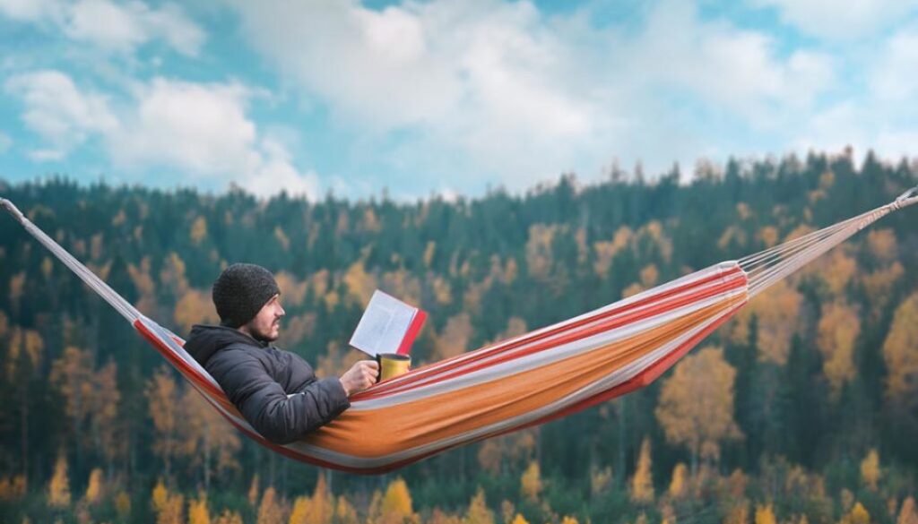 A man sits in a hammock and reads a book in a picturesque place. Mug in his right hand.