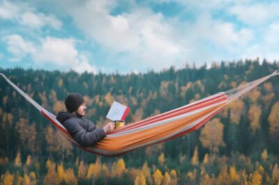 A man sits in a hammock and reads a book in a picturesque place. Mug in his right hand.